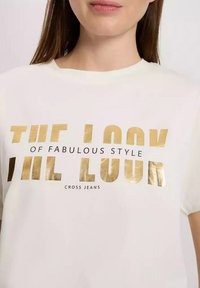 Biała bawełniana koszulka z krótkim rękawem z wyrazistym metalicznym złotym napisem "WYGLĄD FANTASTYCZNEGO STYLU" i "CROSS JEANS."