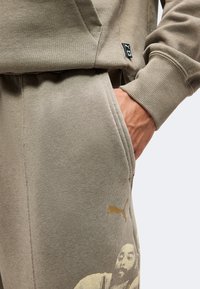 Main dans la poche d’un pantalon de survêtement beige Puma avec un motif visage imprimé, accompagné d’un sweat-shirt beige assorti avec poignets côtelés et petite étiquette logo.