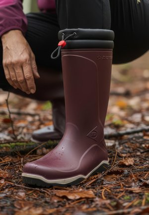 Person wearing purple waterproof boot with black cuff and red toggle, crouching on wet forest floor covered with leaves and twigs.