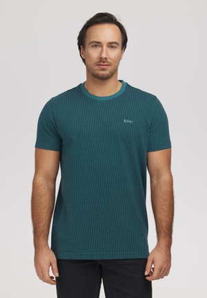 Mężczyzna w morsko niebieskim, teksturowanym t-shircie z krótkim rękawem i czarnych spodniach, stojący na tle jednolitego jasnego tła, zwrócony przodem z neutralnym wyrazem twarzy.