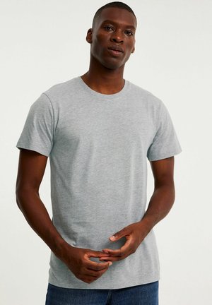 Mann mit kurzen Haaren, trägt ein schlichtes hellgraues T-Shirt und blaue Jeans, steht mit locker verschränkten Händen auf Taillenhöhe.