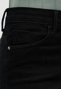 Κοντινό πλάνο στη μέση και την μπροστινή τσέπη μαύρου τζιν με ασημί ούπα, σε συνδυασμό με πράσινο πλεκτό τοπ.