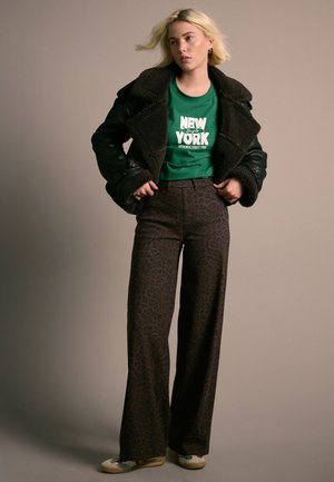 Femme aux cheveux blonds portant une veste en peau retournée noire, un t-shirt vert "New York", un pantalon large imprimé léopard marron et des chaussures beiges, debout devant un fond beige.