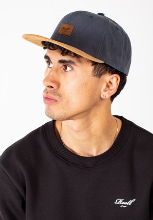 Dark gray cap with a light brown suede brim, featuring a rectangular leather patch on the front, worn with a black sweatshirt.