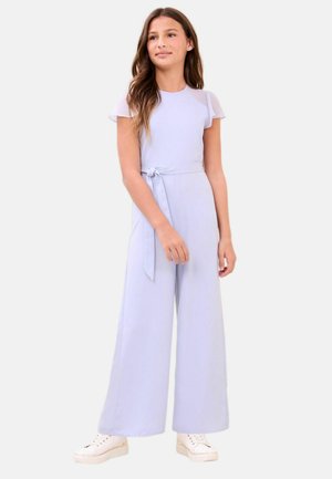 Dziewczyna o długich brązowych włosach nosi jasnofioletowy kombinezon z szerokimi nogawkami, krótkimi rękawami i wiązaniem w talii, stoi w białych trampkach.