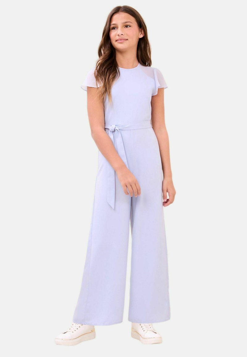 Pikkade pruunide juustega tüdruk kannab helesinist laia säärtega kombinesooni, millel on lühikesed varrukad ja vöökand, ning seisab valgete tossudega.