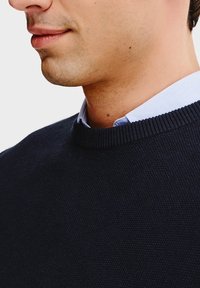 Granatowy sweter dzianinowy z okrągłym dekoltem, strukturalną tkaniną i widocznymi ściągaczami, nałożony na jasnoniebieską koszulę z kołnierzykiem.