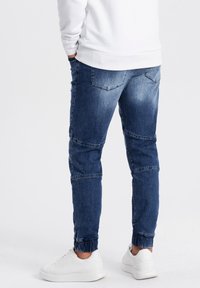 Tummansiniset denimi-joggersit, joissa on kulunut pinta. Ominaisuuksiin kuuluvat kapenevat lahkeet, elastiset hihnat ja takataskut, yhdistettyinä valkoisiin lenkkareihin.