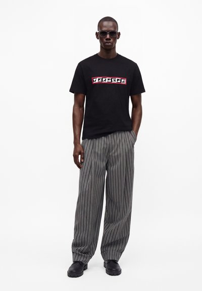 Homme portant un t-shirt noir Versace, pantalon ample gris à rayures verticales, chaussures noires et lunettes de soleil noires, debout avec une main dans la poche.