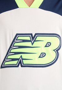 Valkoinen urheilu-paita, jossa on suuri, brodeerattu "NB"-logo tummansinisellä ja neonvihreällä, tekstuuripintaisella kankaalla ja erottuvalla kauluksen suunnittelulla.