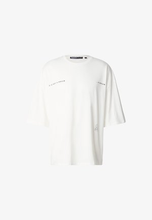 Fehér, túlméretezett pamut póló rövid ujjakkal, fekete "NEM ÁLOM" felirattal és egy minimalista grafikával az alsó jobb oldalon.