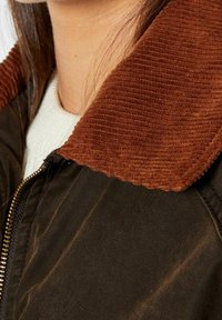 Brun kordfløjlskrage på en mørk jakke, med lynlås lukning og tekstureret stof; har en lysfarvet sweater under.