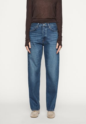 Person iført højtaljede blå jeans med lige ben, en brun langærmet top og beige sneakers, stående mod en ensfarvet baggrund.