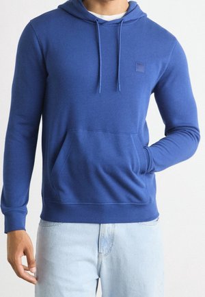 Hombre con sudadera azul con bolsillo delantero y cordones, combinado con jeans azul claro, sobre un fondo blanco liso.