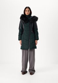 Dark green puffer coat with a faux fur hood, black leather accents on shoulders, and chevron quilted pattern. Zipper closure, knee-length design.