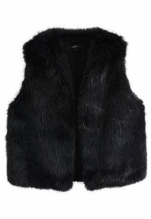 Vrouw met een zwart faux fur vest over een witte trui met lange mouwen, staand tegen een bakstenen muur buiten.
