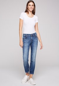 Jeans de mezclilla azul con corte ajustado, acabado desgastado y bolsillos frontales, combinados con una camiseta blanca ajustada y zapatillas blancas.