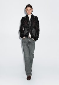 Czarna i szara kurtka zapinana na zamek z wzorem w romby, miękka w dotyku, z prążkowanym kołnierzem, zestawiona z luźnymi szarymi dżinsami i brązowymi butami.