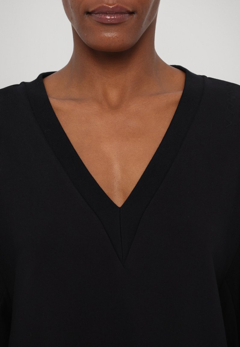 Haut noir à col en V fabriqué en tissu doux, présentant un décolleté profond et des bords nets. Coutures détaillées visibles à l'épaule.