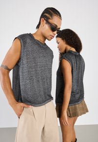 Two models wearing loose-fitting, sleeveless, dark gray tops made of a textured fabric, paired with beige trousers and a plaid skirt.