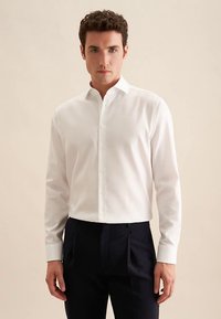 Weißes Langarmhemd aus glattem Stoff, mit spitzem Kragen und durchgehend geknöpftem Vorderteil, kombiniert mit dunklen Hosen.