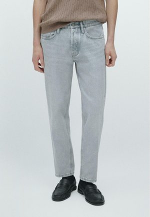 Osoba oblečená v světle šedých džínách s rovnými nohavicemi, černých mokasínech a hnědém pleteném tílku bez rukávů, stojící s uvolněnými pažemi podél těla.