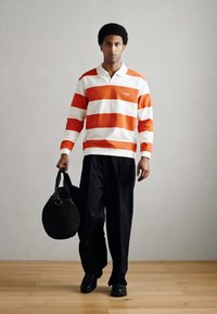 Polo às riscas em laranja e branco, combinado com calças pretas largas. O modelo leva uma mala desportiva preta e calça sapatos pretos.