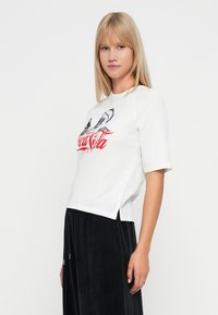 Blondynka ubrana w białą koszulę z grafiką Coca-Cola i luźne czarne spodnie, stojąca na tle czystej białej ściany.