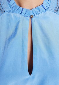 Blouse bleu clair avec un col à volants, comprenant une découpe en forme de larme et un bouton décoratif en haut. Tissu doux et lisse.