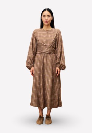 Femme se tient face à l'avant, portant une robe portefeuille à manches longues à carreaux bruns et des chaussures à lacets brunes, contre un fond blanc uni.