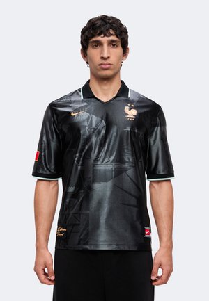 Homme portant le maillot de football noir de la France avec un logo Nike doré et un emblème d'équipe, debout devant un fond clair uni.