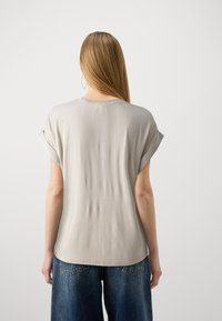 Mujer con cabello largo y liso, vistiendo una camiseta de manga corta gris claro y jeans de mezclilla azul, de espaldas contra un fondo simple.