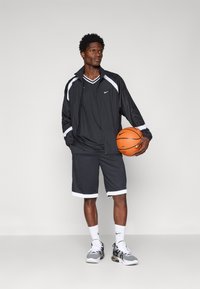 Conjunto deportivo negro de Nike con acentos blancos; incluye una chaqueta con cremallera, camiseta de cuello en V y pantalones cortos. El modelo sostiene una pelota de baloncesto. Zapatillas grises.