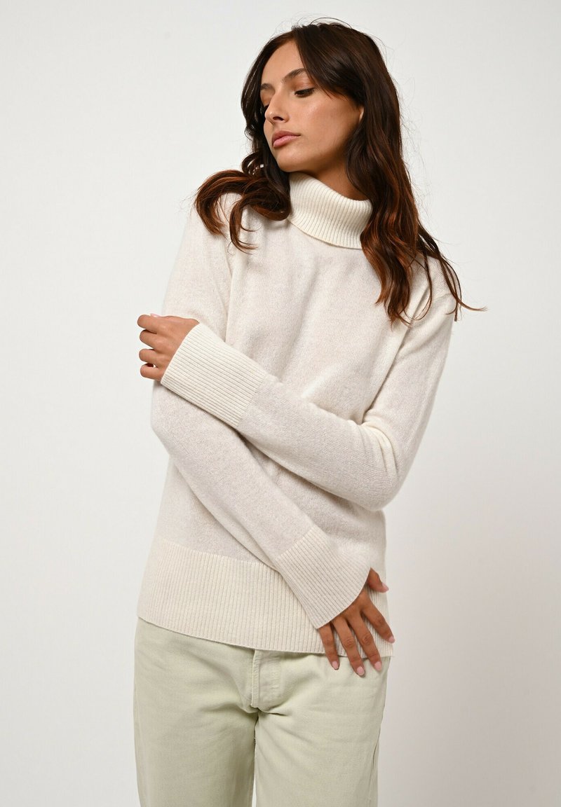 Jasna kremowa sweter turtleneck z prążkowanymi mankietami i dołem. Miękka tekstura, luźny krój i minimalistyczny design. Połączony z jasną zieloną spódnicą.