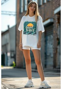 Witte oversized t-shirt met een kleurrijke print van een zonsondergang en golven, gecombineerd met witte shorts en witte sneakers. Stedelijke achtergrond.