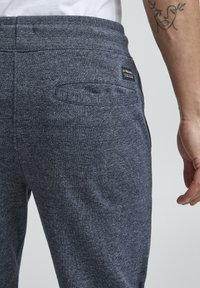 Tumši pelēki, teksturēti sporta bikses ar aizmugurējo kabatu un mazu zīmola birku. Materiāls šķiet mīksts un nedaudz elastīgs.