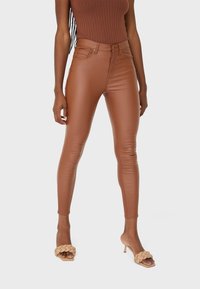 Braune, lederähnliche High-Waist-Skinny-Jeans mit glatter Textur, ausgestattet mit Vordertaschen und einem Knopfverschluss; kombiniert mit beigen Riemchen-High-Heels.