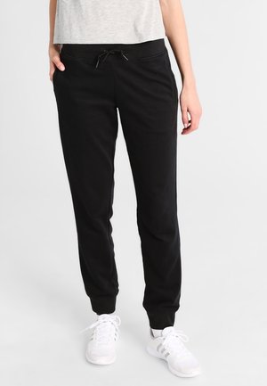 Vrouw draagt zwarte joggerbroek met trekkoord, witte sneakers en lichtgrijze cropped top, staand tegen een effen lichte achtergrond.