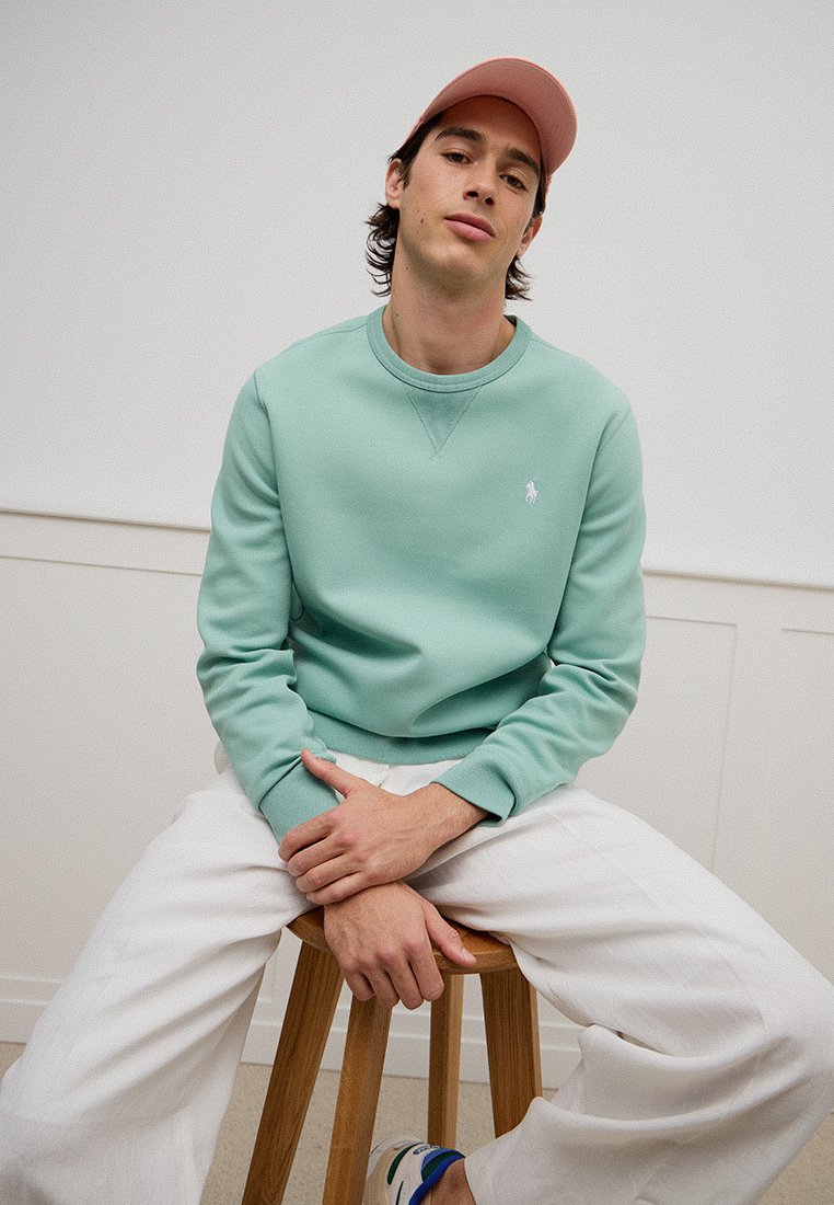 Helle grüne Sweatshirt mit Rundhalsausschnitt, ausgestattet mit einem kleinen weißen Logo. Kombiniert mit weißen Hosen und einer pinken Kappe. Sitzt auf einem hölzernen Hocker.