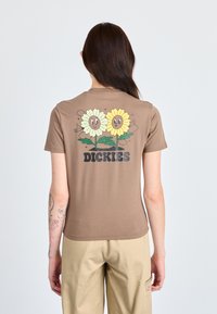 Ruskea t-paita, jossa on kuvio kahdesta hymyilevästä auringonkukasta ruskeilla keskiosilla, ja sanan "DICKIES" painatus alla. Lyhyet hihat ja muotoiltu malli.