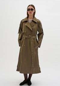 Lungo trench coat beige con vita cinturata, chiusura a bottoni, due tasche laterali e spalline. Indossato sopra collant neri e scarpe.