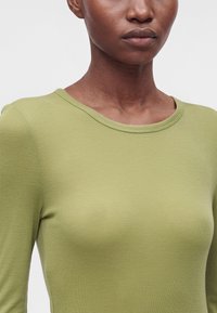 Gros plan d'une personne portant une chemise vert clair à manches longues avec un col rond sur un fond clair uni.