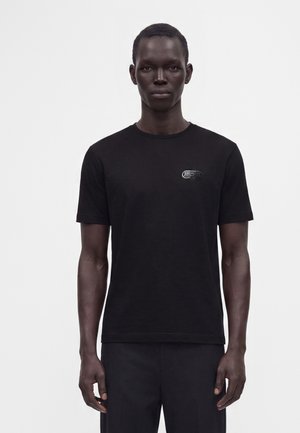 Άνδρας που φοράει απλό μαύρο κοντομάνικο T-shirt με μικρό μαύρο λογότυπο στο αριστερό στήθος, στέκεται μπροστά σε απλό λευκό φόντο.