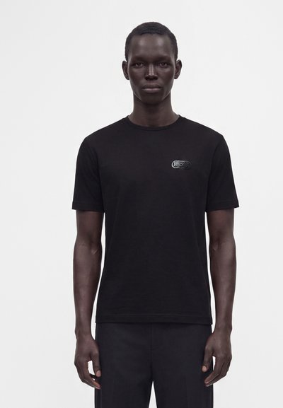 Άνδρας που φοράει απλό μαύρο κοντομάνικο T-shirt με μικρό μαύρο λογότυπο στο αριστερό στήθος, στέκεται μπροστά σε απλό λευκό φόντο.