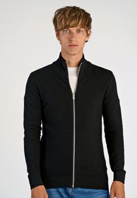 Svart stickad zip-up-tröja med hög krage, med ett texturerat mönster och ribbade muddar. Dragkedjan går ner över framsidan.