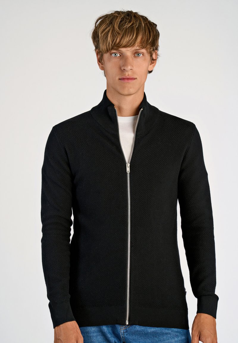 Svart stickad zip-up-tröja med hög krage, med ett texturerat mönster och ribbade muddar. Dragkedjan går ner över framsidan.