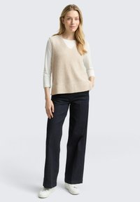 Beige gebreide mouwloze trui met een V-hals, gedragen over een witte blouse met lange mouwen. Gecombineerd met donkere wijde jeans en witte sneakers.
