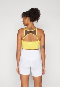 Crop top de sport jaune avec un dos en mesh, associé à un short blanc taille haute. Marque notable au dos du top. Texture lisse.