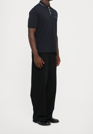 Man wearing a navy short-sleeve polo shirt with white piping, black wide-leg trousers, and black shoes, standing against a white background.