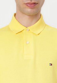 Cămașă polo galbenă, realizată dintr-un material texturat, cu guler clasic, având două butoane și un mic logo în culorile roșu, alb și albastru pe piept.